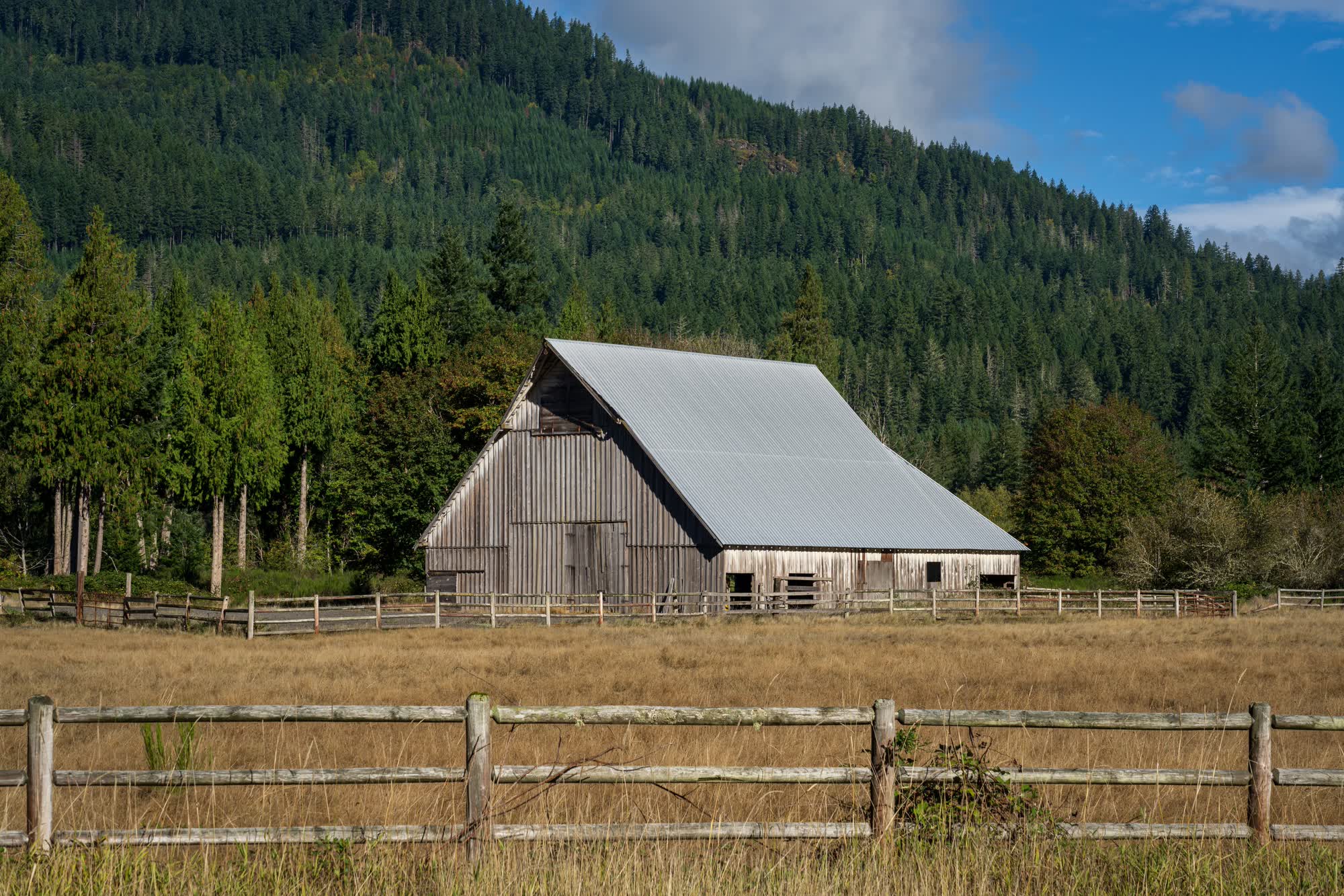 A weathered grey barn and a rail fence below a forested hillside